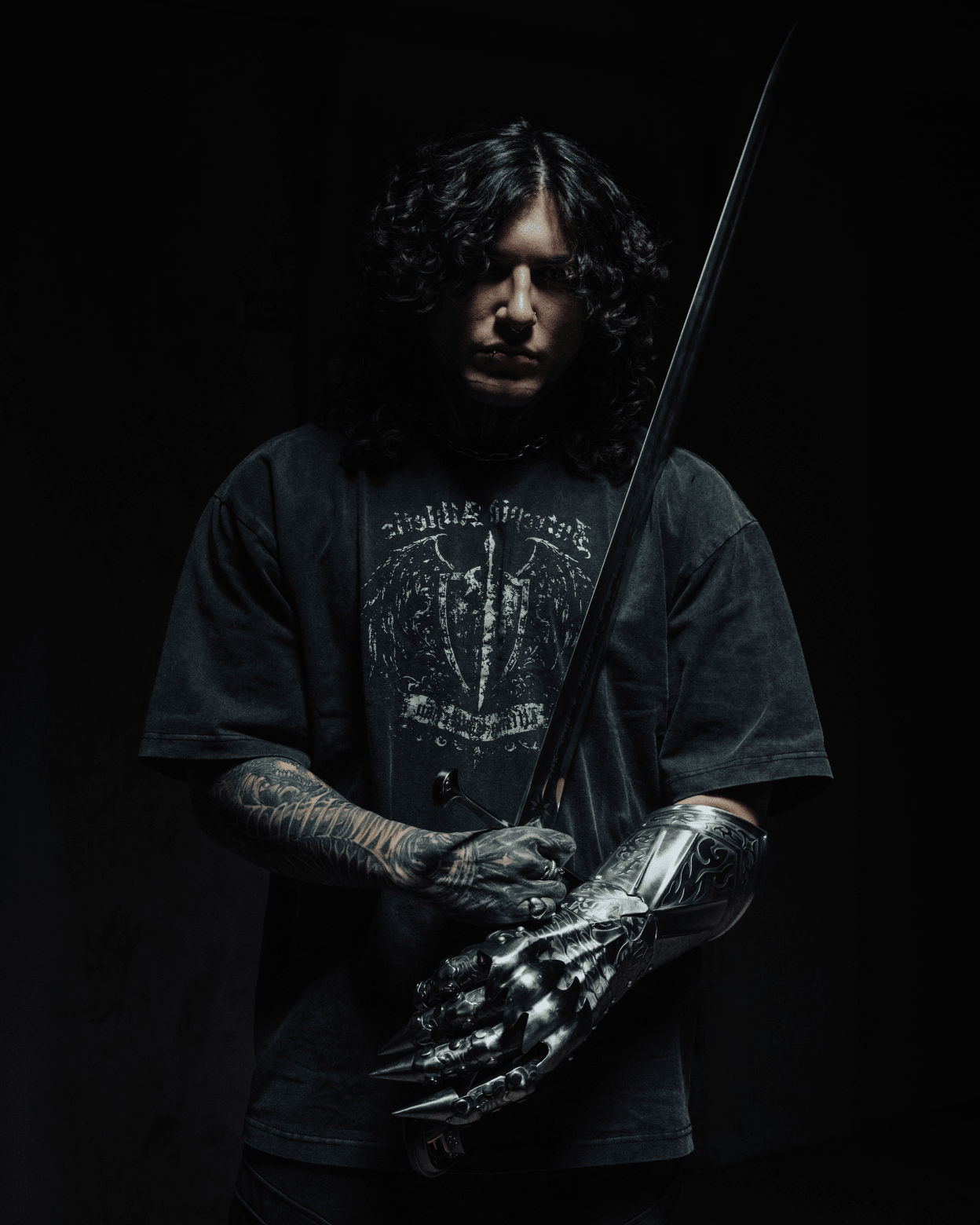 Person wearing a black t-shirt with a graphic design, holding a sword against a dark background
