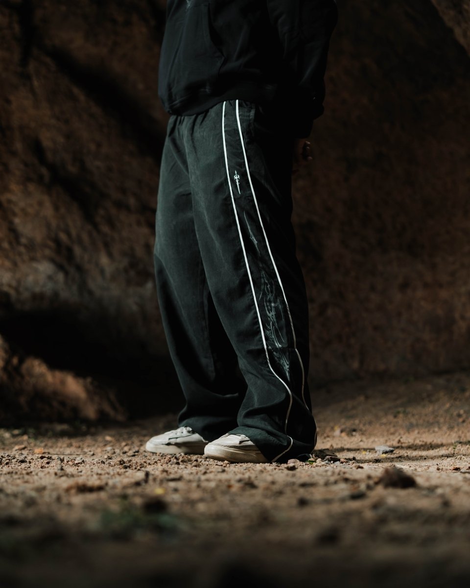 Person wearing dark green pants with white stripes standing on a dirt ground.