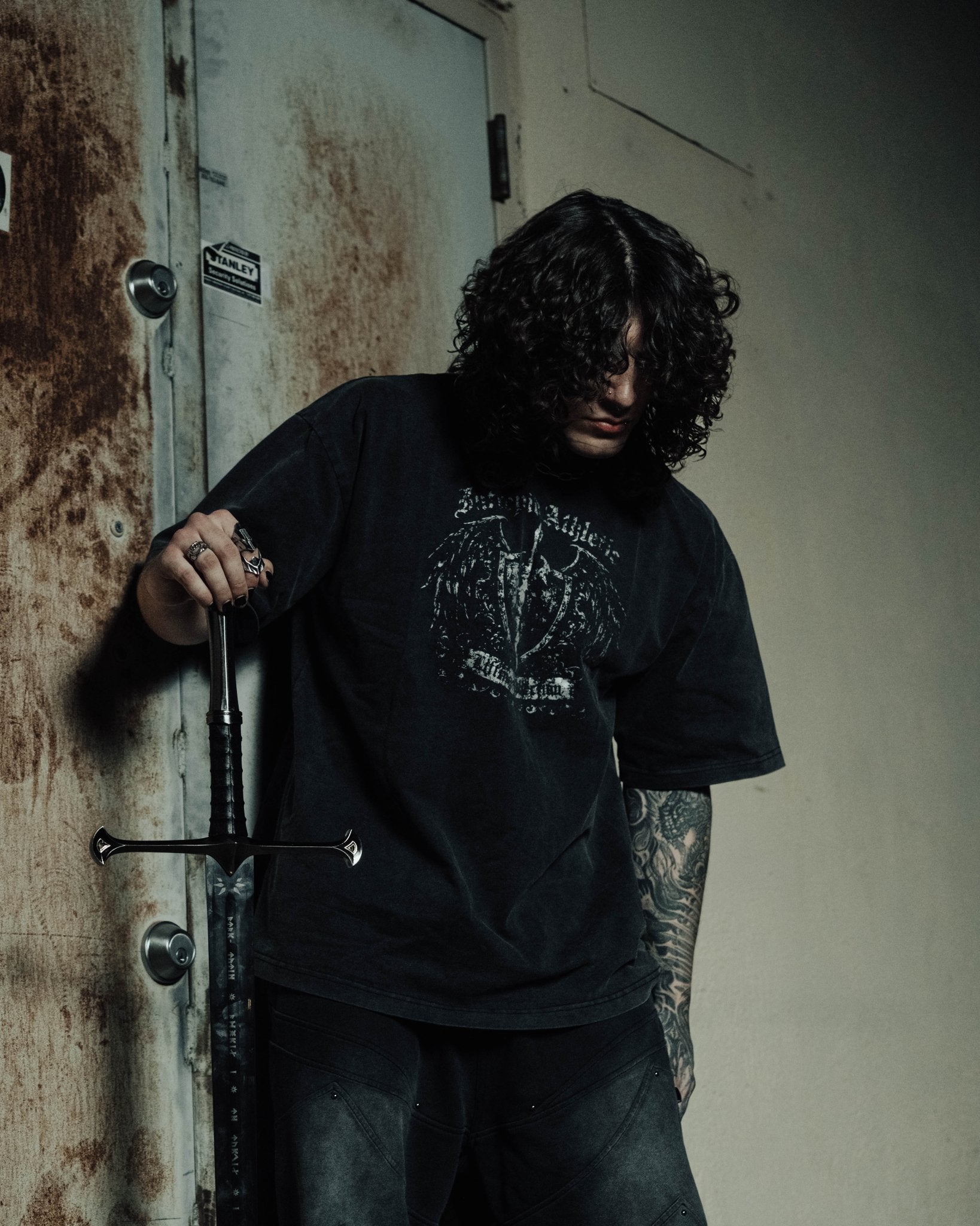 Person wearing a black t-shirt with a graphic design, leaning against a textured wall.