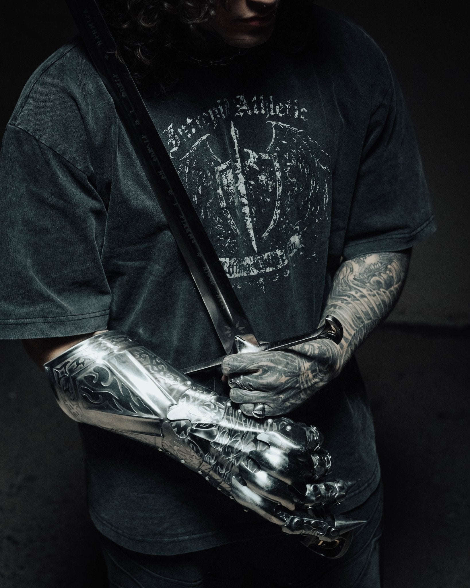 Person wearing a dark t-shirt with a graphic design, holding a large knife, and wearing metallic arm guards against a dark background.