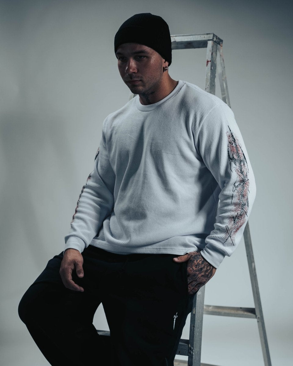 Man wearing a light gray sweater with tattoos on his arms, standing next to a ladder against a plain background.