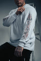 Person wearing a light gray sweater with intricate designs on sleeves and chest, standing against a neutral background.