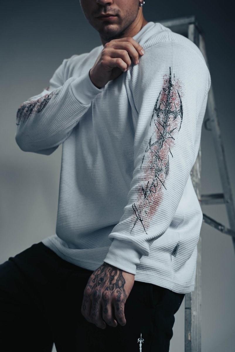 Person wearing a light gray sweater with intricate designs on sleeves and chest, standing against a neutral background.