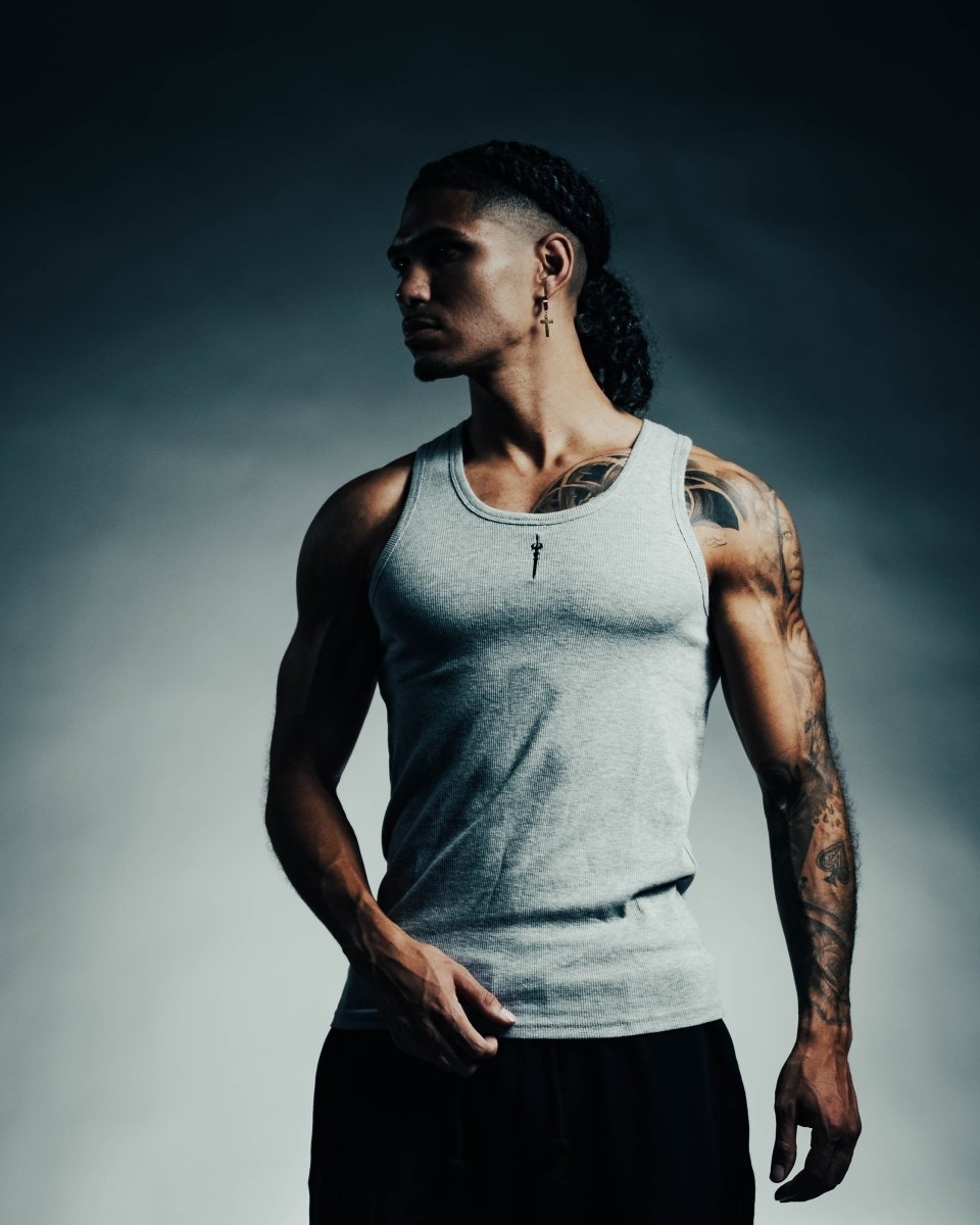 Man wearing a gray tank top with tattoos on his arms against a dark background