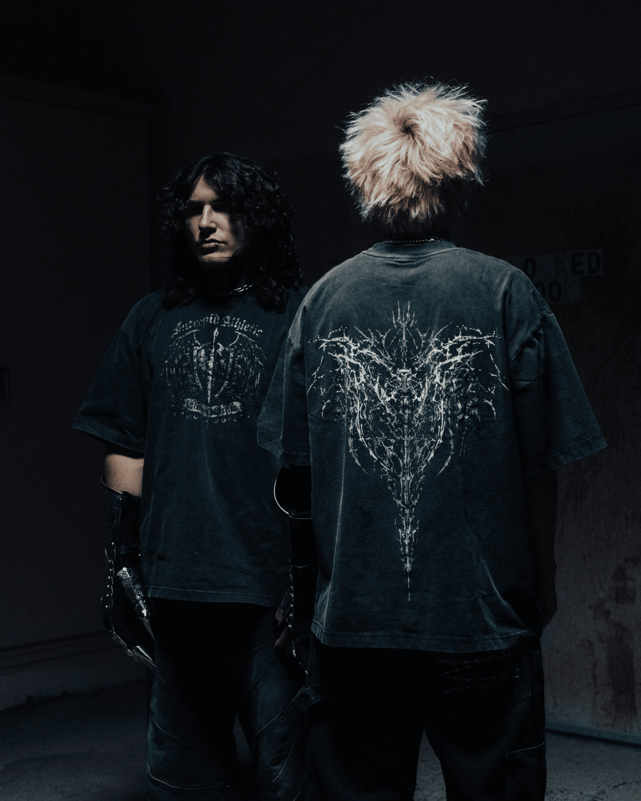 Two people wearing dark t-shirts with unique designs, one facing the camera and the other with back to the camera.